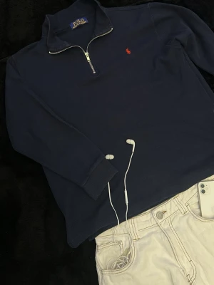 Marinblå halvzip från Polo Ralph Lauren - Klassisk marinblå långärmad tröja från Polo Ralph Lauren med halvzip och liten röd logga broderad på bröstet. Tröjan har hög krage och ribbade muddar vid ärmslut och nederkant. Perfekt för en stilren och avslappnad look.