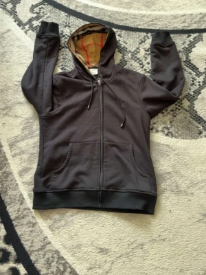 Svart hoodie från Burberry - Säljer en svart hoodie från Burberry med dragkedja, fickor framtill och klassiskt rutigt foder i huvan. Broderad Burberry-logga på bröstet och justerbara snören i huvan. Perfekt för dig som gillar stilrena och exklusiva plagg.