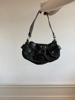 Svart axelremsväska  - Cool svart handväska i croc-mönstrat konstläder med bred axelrem och silverfärgade metalldetaljer. Väskan har ett stort spänne framtill och dekorativa remmar på sidorna. Perfekt accessoar