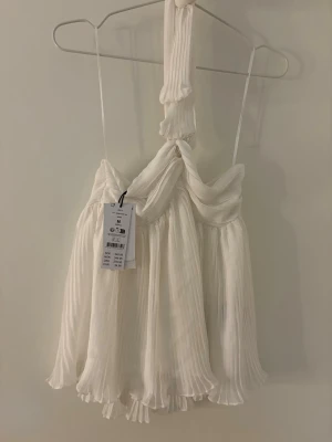 Vit halterneck topp - Superfin vit halterneck topp med plisserat tyg och flowig passform. Toppen har tunna axelband och en snygg drapering framtill. Perfekt för dig som vill ha en luftig och trendig look i sommar.