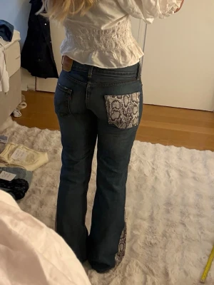 Blå jeans med ormmönstrade detaljer - Unika blå jeans med utsvängda ben och coola ormmönstrade detaljer på bakficka, ficka fram och längs bensluten. Snygg kontrast mellan denim och det gråa ormmönstret. Sytt på ormmönstret själv.