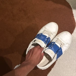 Valentino open  - Säljer ett par vita sneakers från Valentino med bred blå rem över vristen och coola nitar på hälen. Skorna har snörning och är tillverkade i skinn för en lyxig känsla. Perfekta för dig som vill ha något unikt och stilrent.