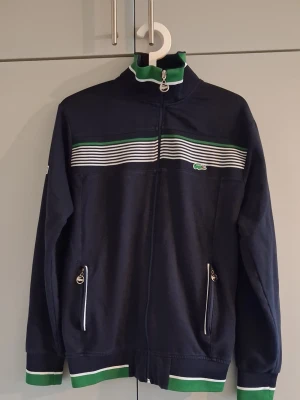 Marinblå ziptröja från Lacoste set - Snygg marinblå ziptröja från Lacoste med vita och gröna detaljer. Tröjan har dragkedja framtill, två fickor med dragkedja och randigt mönster över bröstet. Ribbad mudd i ärmslut och nederkant med gröna och vita ränder. Perfekt för en sportig look.