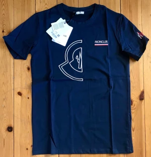 Mörkblå Moncler t-shirt med logga - Snygg mörkblå t-shirt från Moncler med vit broderad logga på bröstet och diskret Moncler-tryck på ärmen. Klassisk rund hals och korta ärmar. Tillverkad i mjuk bomull som känns skön mot huden. Perfekt för en stilren och trendig look.