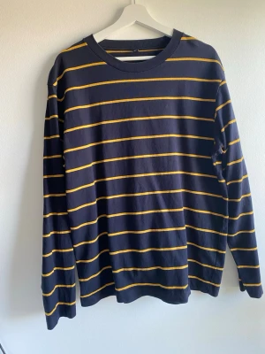 Randig långärmad tröja från Uniqlo L - Snygg långärmad tröja från Uniqlo i mörkblått med tunna gula ränder. Tröjan har en klassisk rund halsringning och är gjord i mjuk bomull. Perfekt för en avslappnad och stilren look.