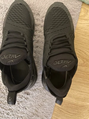 Nike Air Max 270 helsvarta sneakers - Säljer ett par helsvarta Nike Air Max 270 sneakers med mesh-ovandel och synlig Air Max-dämpning i hälen. Skorna har en sportig design, snörning framtill och diskreta loggor på plös och sidor. Perfekta för dig som gillar stilrena och bekväma sneakers.