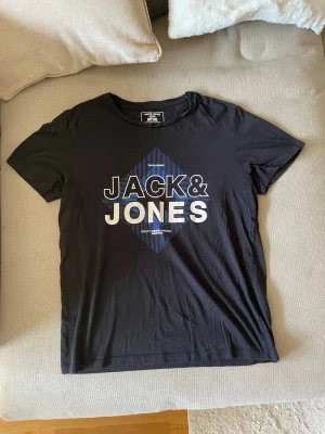 Svart Jack & Jones t-shirt - Svart t-shirt från Jack & Jones med tryck i vitt och blått framtill. Klassisk rund hals och korta ärmar. Perfekt för en avslappnad och stilren look.