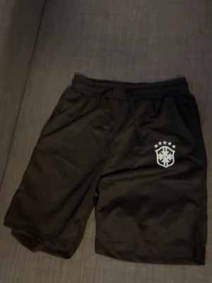 Svarta shorts med tryck och snörning - Svarta shorts med vitt tryck på ena benet och snörning i midjan. De har en sportig look och är perfekta för träning eller chill. Materialet känns som polyester och de har en bekväm passform.