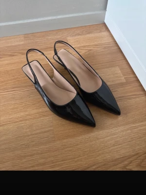 Svarta lackpumps med låg klack - Snygga svarta pumps med spetsig tå och låg klack. Skorna har öppen häl med smal rem runt ankeln och är tillverkade i glansigt syntetmaterial som ger en elegant look. Perfekta för dig som gillar stilrena och trendiga skor. 