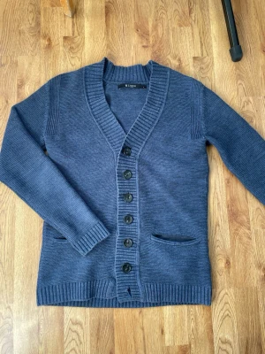 Marinblå cardigan Tiger of Sweden - Snygg marinblå kofta från Tiger of Sweden med djup V-ringning och knäppning framtill. Koftan har två fickor, ribbade muddar och är stickad i ett mjukt material. Perfekt för lager-på-lager och stilren look.