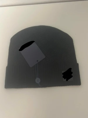 Svart ribbstickad mössa - Snygg svart mössa med ribbstickad kant och diskret logotypmärke framtill. Klassisk form som passar till de flesta stilar. Perfekt accessoar för kyliga dagar och enkel att matcha med olika outfits.