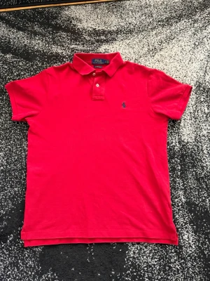 Röd pikétröja från Polo Ralph Lauren - Klassisk röd pikétröja från Polo Ralph Lauren med blå broderad logga på bröstet. Tröjan har korta ärmar, krage och knappar vid halsen. Tillverkad i bomull för en bekväm och luftig känsla.