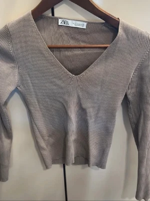 Beige ribbad tröja från Zara - Ribbstickad beige tröja från Zara med djup V-ringning och långa ärmar. Tröjan är figurnära och har en mjuk, stretchig känsla som passar perfekt till lager-på-lager-stil. Enkel och stilren design som är lätt att matcha.