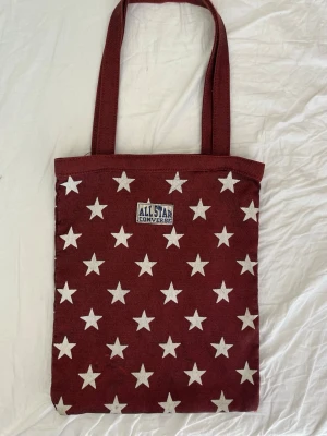 Röd axelväska med stjärnor, Converse - Snygg röd totebag från Converse med vita stjärnor över hela väskan och klassisk All Star-logga framtill. Väskan har två långa handtag och är tillverkad i slitstarkt tygmaterial. Perfekt för att bära böcker, laptop eller shopping. Alla bud över 125kr godkänns!