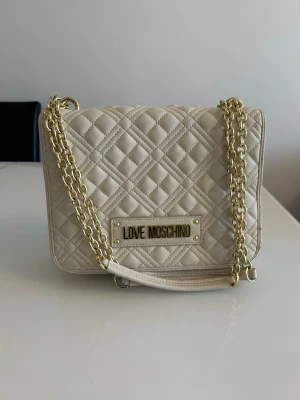 Beige quiltad axelväska Love Moschino - Snygg beige axelväska från Love Moschino med quiltat mönster och guldfärgad kedjerem. Väskan har en rektangulär form och en metallskylt med loggan på framsidan. Tillverkad i konstläder som ger en lyxig känsla.