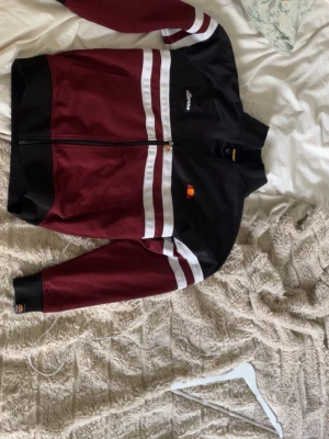 Ellesse Rimini track jacket vinröd/svart - Snygg track jacket från Ellesse, modell Rimini, i vinrött och svart med vita och röda ränder över bröstet och ärmarna. Dragkedja framtill, hög krage och broderad logga på bröstet. Tillverkad i ett glansigt syntetmaterial som ger en retro sportig look.
