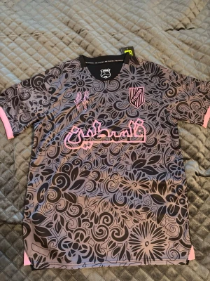 FC Palestina matchtröja blommig grå/rosa - Unik matchtröja från FC Palestina i storlek Large. Tröjan har ett grått och svart blommönster med rosa detaljer, logga och text på bröstet. Tillverkad i lätt och snabbtorkande material, perfekt för fotboll eller träning. Rund hals och korta ärmar.