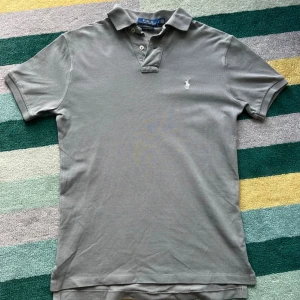 Grå pikétröja från Ralph Lauren - Klassisk grå pikétröja från Ralph Lauren med vit broderad logga på bröstet. Tröjan har korta ärmar, krage och knappar vid halsen. Tillverkad i bomull för en bekväm och stilren look. Plagget är custom slimfit så den sitter perfekt!