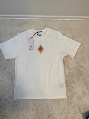 Vit t-shirt från C.P. Company - Snygg vit t-shirt från C.P. Company med en liten brun logga på bröstet. Klassisk rund halsringning och korta ärmar. Tillverkad i mjuk bomull som känns skön mot huden. Perfekt för en clean och stilren look. Passar även L