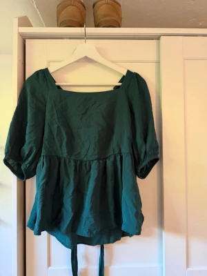 Grön blus med knyt och puffärm - Snygg grön blus med puffiga trekvartsärmar och två knytband i ryggen. Blusen har en lös passform och fyrkantig ringning framtill. Perfekt för dig som gillar en avslappnad men trendig stil.