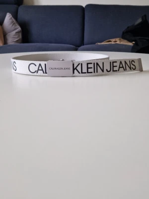 Calvin Klein bälte - Snyggt vitt bälte från Calvin Klein Jeans med svart logotyptext längs hela bandet. Bältet har ett metallspänne med märkets namn tryckt på. Perfekt accessoar för att lyfta din outfit med en clean och modern vibe.