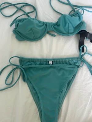 Turkos bikini med knyt - Säljer en snygg turkos bikini med knyt i sidorna på både överdel och underdel. Bikinin har en enkel och stilren design med band som man kan knyta hur man vill. Perfekt för strandhäng eller poolen i sommar! Har tvättat den men inte använt. Från NAKD, storlek 70B för överdelen och S för underdelen. 