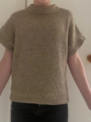 Beige stickad t-shirt med kort ärm - Säljer en beige stickad t-shirt med korta ärmar och ribbad halsringning. Tröjan har en rak passform och är gjord i ett mjukt, ull-liknande material. Perfekt för lager på lager och enkel att styla till olika outfits.