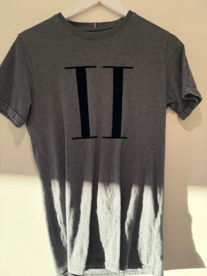 Grå t-shirt från Les Deux - Snygg grå t-shirt från Les Deux med stort svart tryck av två romerska ettor på bröstet. Klassisk rund halsringning och korta ärmar. Tillverkad i mjuk bomull som känns skön mot huden. Perfekt till jeans eller shorts.