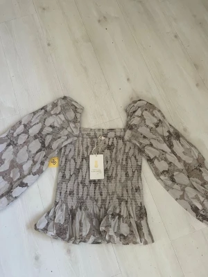 Mönstrad blus från Pernilla Wahlgren - Trendig blus i beige och brunt mönster från Pernilla Wahlgren. Blusen har smockad kropp, volang nertill och vida, transparenta långärmar. Perfekt för dig som gillar en romantisk och bohemisk stil. Materialet är lätt och luftigt.