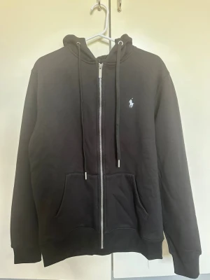 Svart zip-hoodie från Ralph Lauren - Svart hoodie med dragkedja från Ralph Lauren. Klassisk design med huva, dragsnören och känguruficka framtill. Liten vit logga broderad på bröstet. Perfekt för en avslappnad och stilren look.