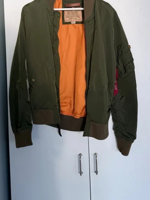 Grön bomberjacka från Alpha Industries - Stilren grön bomberjacka från Alpha Industries med orange foder och klassisk 'Remove Before Flight'-tag på ärmen. Jackan har ribbade muddar, dragkedja och flera fickor. Perfekt för dig som gillar streetwear och tidlös design.