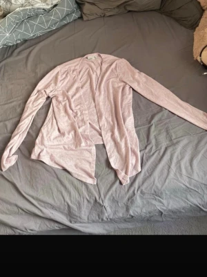 Ljusrosa öppen kofta - Säljer en ljusrosa, tunn och öppen kofta med lång ärm. Perfekt att slänga över en t-shirt eller linne för en chill och avslappnad look. Koftan har ingen knäppning och är i ett mjukt, stretchigt material.