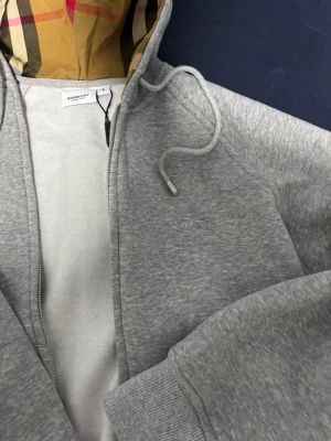 Grå zip hoodie från Burberry - Snygg grå zip hoodie från Burberry med klassiskt rutigt foder i huvan och diskret broderad logga på bröstet. Hoodien har dragsko i huvan, fickor framtill och ribbade muddar. Perfekt för en avslappnad och stilren look.