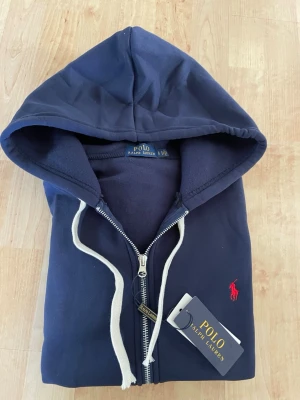 Marinblå hoodie från Polo Ralph Lauren - Snygg marinblå hoodie från Polo Ralph Lauren med dragkedja, vit dragsko och klassisk röd logga broderad på bröstet. Tillverkad i mjukt bomullsmaterial och har en stor huva. Perfekt för en avslappnad och stilren look.