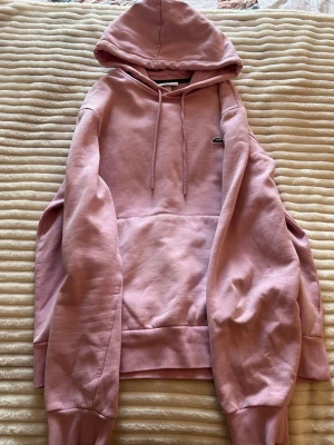 Lacoste hoodie  - Säljer en ljusrosa hoodie från Lacoste med klassisk krokodillogga på bröstet. Hoodien har huva med dragsko och en stor magficka. Tillverkad i mjukt bomullsmaterial, perfekt för en chill och avslappnad stil.