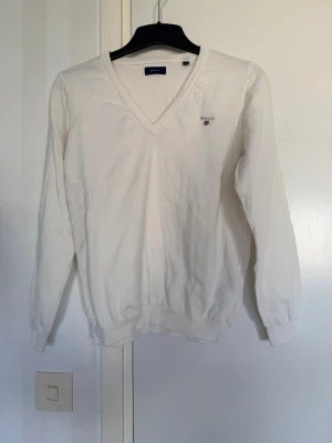 Vit stickad tröja från GANT - Vit stickad tröja från GANT med V-ringning och broderad logga på bröstet. Tröjan har långa ärmar och ribbade muddar vid ärmslut och nederkant. Perfekt för en clean och klassisk stil.
