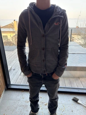 Mörkgrå hoodie från Hollister - Snygg mörkgrå hoodie från Hollister med dragkedja och knappar framtill. Stor logga på ärmen och liten logga på bröstet. Utrustad med huva och vita dragsnören samt två fickor framtill. Perfekt för en avslappnad och trendig stil. Passar 150 - 165 cm