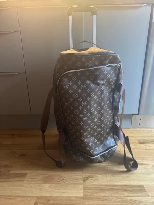  brun resväska på hjul  - Säljer en brun resväska. Väskan har två bärhandtag, axelrem och utdragbart handtag i metall. Tillverkad i canvas med detaljer i brunt och guldiga dragkedjor. Perfekt för weekendresor och har en rymlig form.