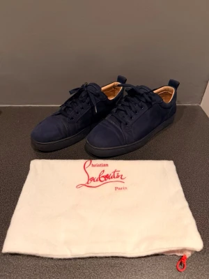 Mörkblå sneakers från Louboutin - Snygga mörkblå sneakers från Christian Louboutin i mocka med svarta sulor och matchande mörkblå skosnören. Skorna har en klassisk låg profil och beige innersida. Perfekta för dig som vill ha en stilren och exklusiv look.