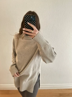 Beige oversized stickad polotröja - Mysig beige stickad polotröja med oversized passform och breda ribbade muddar vid ärmslut och nederkant. Tröjan har slitsar i sidorna och är perfekt för lager-på-lager-stil. Materialet ser ut att vara mjukt och värmande, perfekt för kyliga dagar.