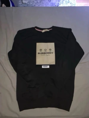 Svart Burberry sweatshirt med beige patch - Cool svart sweatshirt från Burberry med stor beige patch på bröstet och tydlig logga. Tröjan har rund halsringning och ribbade muddar. Perfekt för dig som gillar streetwear och vill sticka ut med ett exklusivt plagg.