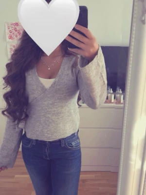 Grå omlottstickad tröja - Mysig grå stickad omlott-tröja med knytning i sidan och långa ärmar. Tröjan har en djup v-ringning och ribbstickade muddar. Perfekt att matcha med både jeans och kjol för en avslappnad look.