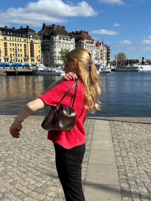 Mörkbrun handväska från Gina Tricot - Säljer en stilren mörkbrun handväska i slätt material med långa dubbla axelband. Väskan har en avlång, mjukt rektangulär form och en enkel ficka framtill. Perfekt för dig som gillar minimalistisk och klassisk design.