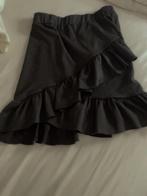 Svart volangkjol från NLY Trend - Säljer en svart kortkjol från NLY Trend med bred resår i midjan och snygga volangdetaljer. Kjolen har omlottkänsla och är gjord i ett mjukt, lätt material som ger en skön passform. Perfekt för dig som gillar trendiga och feminina plagg.