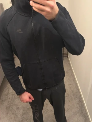 Svart Nike zip-hoodie - Svart hoodie från Nike med dragkedja hela vägen, diskret Nike-logga på bröstet och två stora fickor framtill. Klassisk sportig look med hög krage och mjukt material, perfekt för chill eller träning.