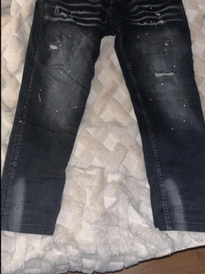 Dsquared2 jeans  - Säljer ett par svarta jeans från Dsquared2 med coola slitningar och färgstänk. Jeansen har en smal passform och tydliga slitdetaljer framtill. Bakfickan har Dsquared2-logga i rött. Perfekta för en trendig streetstyle-look.