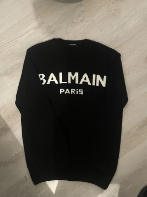  Balmain Tröja  - Säljer en svart stickad tröja från Balmain Paris med vit logga framtill. Tröjan har rund halsringning och ribbade muddar vid halsen. Perfekt för dig som gillar exklusiva streetwear-plagg och vill sticka ut med en ikonisk logga.