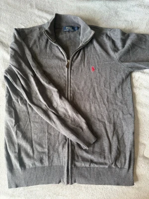 Grå ziptröja från Polo Ralph Lauren - Snygg grå ziptröja från Polo Ralph Lauren med klassisk krage och dragkedja framtill. Tröjan har ribbade muddar och nederkant samt en ikonisk rosa logga broderad på bröstet. Perfekt för en stilren och avslappnad look.