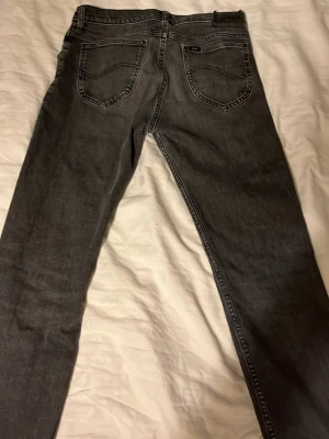 Grå Lee jeans - Säljer ett par grå Luke jeans från Lee med klassisk femficksdesign och svarta sömmar. Jeansen har en något tvättad look och raka ben. Perfekta för dig som gillar en stilren och avslappnad vibe.