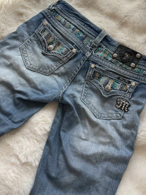 Miss Me ljusblå jeans med strass - Säljer ett par ljusblå jeans från Miss Me med snygga slitningar och detaljerade bakfickor med strass och turkosa pärlor. Jeansen har silverfärgade knappar och tydliga kontrastsömmar. Perfekta för dig som gillar statement-jeans med unika detaljer.